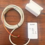 White electronic module and a long coiled cable with colorful wires on a wooden table (Mitsubishi Electric device visible).